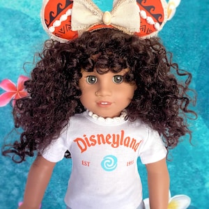 K&ouml;nnte beinhalten: Puppe mit einem wei&szlig;en T-Shirt mit orangefarbenem "Disneyland"-Aufdruck und hellblauen Jeans. Die Puppe hat braune Locken und tr&auml;gt ein Stirnband mit roten und wei&szlig;en Mickey Mouse Ohren. Das Stirnband hat eine beige Schleife.