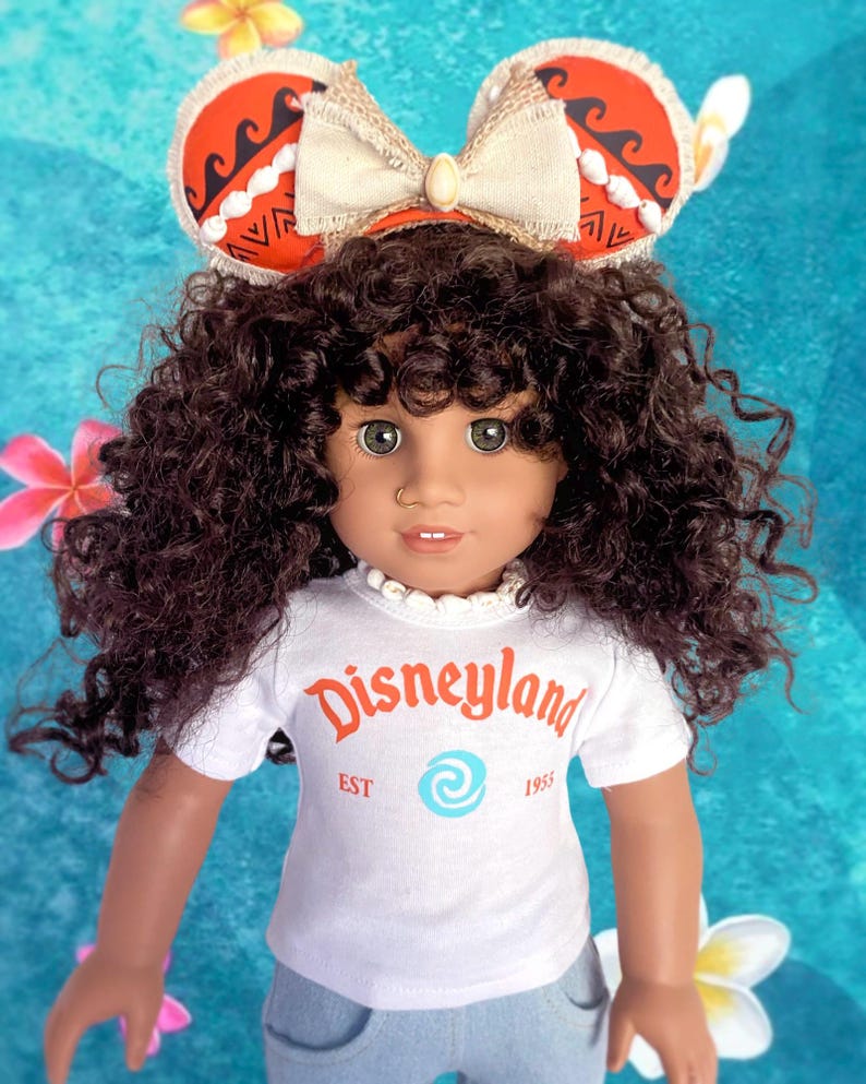K&ouml;nnte beinhalten: Puppe mit einem wei&szlig;en T-Shirt mit orangefarbenem "Disneyland"-Aufdruck und hellblauen Jeans. Die Puppe hat braune Locken und tr&auml;gt ein Stirnband mit roten und wei&szlig;en Mickey Mouse Ohren. Das Stirnband hat eine beige Schleife.