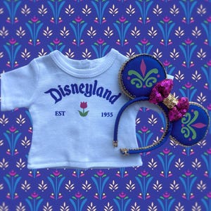 Op de afbeelding: Een wit t-shirt met het woord "Disneyland" in blauw schrift, een rode tulp en het jaar "1955". Een blauwe hoofdband met Mickey Mouse oren, versierd met groene en roze accenten en een roze pailletten strik.