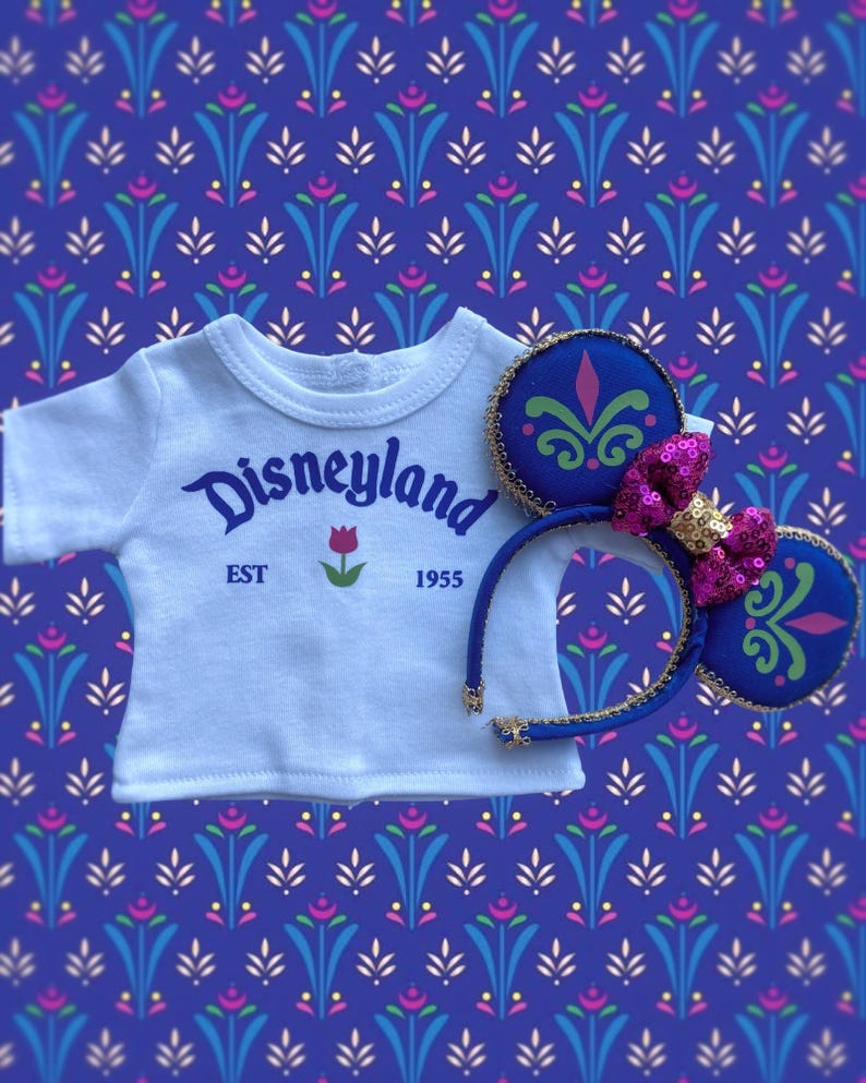 Op de afbeelding: Een wit t-shirt met het woord "Disneyland" in blauw schrift, een rode tulp en het jaar "1955". Een blauwe hoofdband met Mickey Mouse oren, versierd met groene en roze accenten en een roze pailletten strik.