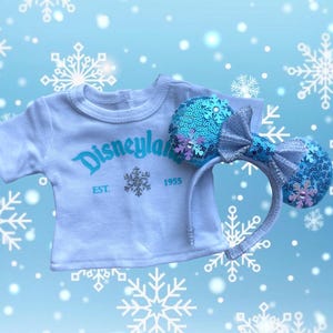 Puede incluir: Una camiseta blanca con "Disneyland" en escritura turquesa y un dise&ntilde;o de copo de nieve, combinada con una diadema de orejas azul con lentejuelas y un lazo plateado. La camiseta tiene "EST. 1955" debajo del logo. Fondo azul claro con copos.