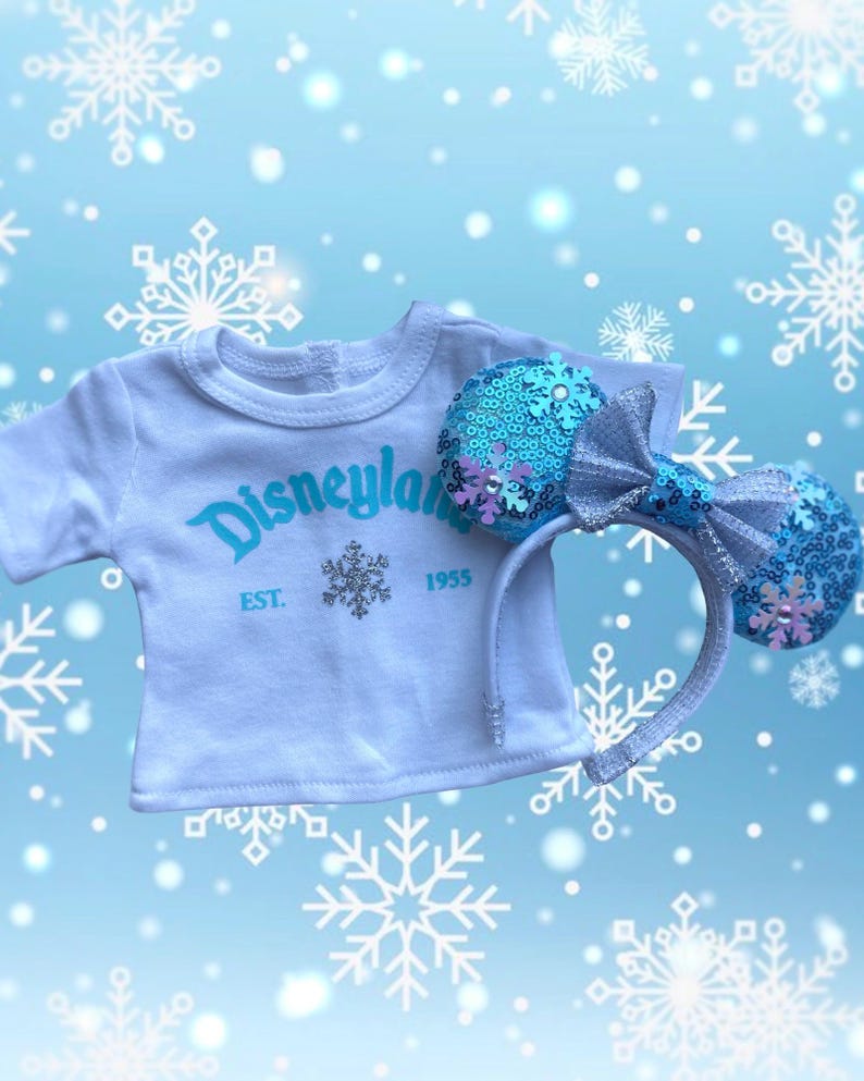 Puede incluir: Una camiseta blanca con "Disneyland" en escritura turquesa y un dise&ntilde;o de copo de nieve, combinada con una diadema de orejas azul con lentejuelas y un lazo plateado. La camiseta tiene "EST. 1955" debajo del logo. Fondo azul claro con copos.