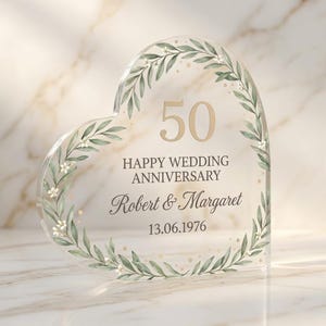Op de afbeelding: Transparante, hartvormige jubileumplaquette met een bloemenkransontwerp. De plaquette toont het gouden getal "50", de tekst "HAPPY WEDDING ANNIVERSARY", de namen "Robert & Margaret" en de datum "13.06.1976".