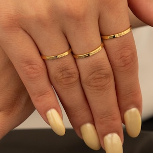 Puede incluir: Una colección de anillos dorados. Los anillos son bandas finas, algunas con patrones grabados y otras con pequeñas inscripciones. Los anillos se muestran en los dedos de una persona, con un fondo oscuro.