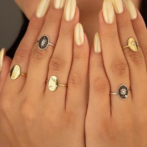 May include: A collection of gold and silver rings with various oval and circular designs. Some rings feature black enamel with starburst or geometric patterns. The rings are displayed on a person's fingers, showcasing the jewelry.
