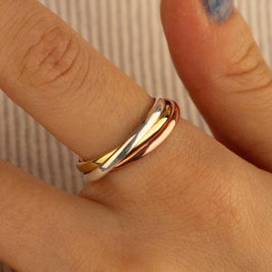 Peut inclure: Une bague à trois anneaux avec des bandes dorées, argentées et or rose. La bague est portée au doigt, mettant en valeur le design entrelacé et les couleurs métalliques. Les bandes sont lisses et polies.