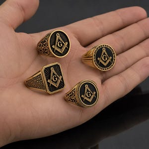 Peut inclure: Quatre bagues dorées avec des symboles maçonniques noirs sont présentées. Les bagues ont différentes formes : rondes, ovales et rectangulaires. Le symbole maçonnique comprend un compas et un carré avec la lettre "G" au centre. Les bagues sont sur une main.