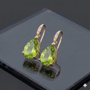 Puede incluir: Un par de pendientes colgantes dorados con piedras preciosas verdes claras en forma de pera. Los pendientes tienen una pequeña fila de piedras transparentes cerca de la parte superior del gancho. Los pendientes se exhiben sobre una superficie reflectante.