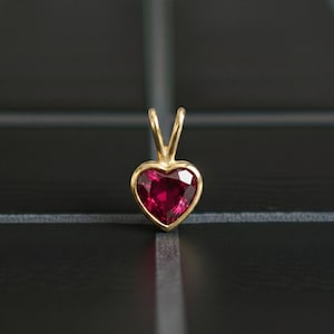 May include: A heart-shaped pendant featuring a vivid red gemstone, set within a gold frame. The pendant has a simple, elegant design with a polished finish. The gemstone is a deep red hue, and the gold setting adds a touch of luxury and sophistication.