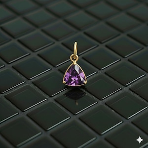 May include: A gold pendant featuring a triangular, purple gemstone. The pendant hangs from a small gold loop. The background is a dark, glossy, tiled surface.
