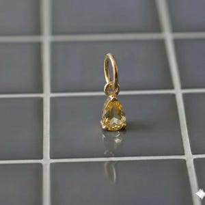 Op de afbeelding: Een gouden hanger met een gefacetteerde, peervormige gele edelsteen die aan een kleine ronde gouden ring hangt. Het sieraad heeft een heldere, doorschijnende gele kleur met een puntige onderkant en een glanzende, gepolijste metalen afwerking.