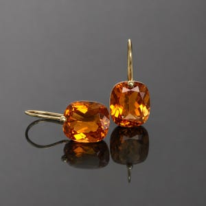 Puede incluir: Un par de pendientes colgantes con piedras preciosas naranjas de talla cojín engastadas en oro. Un pendiente está en posición vertical mientras que el otro está plano, ambos muestran superficies facetadas que captan la luz con ganchos dorados.