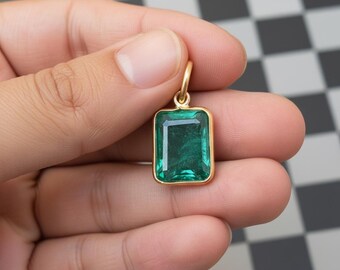 May include: A gold pendant with a rectangular, emerald-green gemstone. The gemstone is set in a gold frame with a small loop at the top for a chain. The pendant is held in a hand against a black and white checkered background.