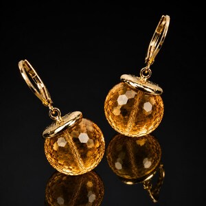 May include: A pair of gold leverback earrings featuring spherical, faceted yellow citrine gemstones. Each earring has a polished gold cap sitting atop the round, textured stone, creating a reflective and bright appearance against a dark background.