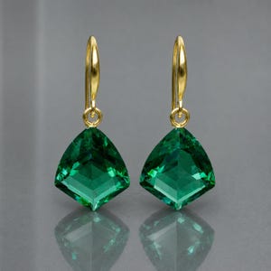 Puede incluir: Un par de pendientes colgantes con piedras de color verde esmeralda talladas en forma triangular, suspendidas de ganchos de metal dorado pulido. Las piedras muestran un tono verde intenso con bordes geométricos definidos que reflejan la luz.