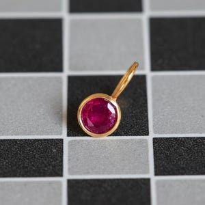 May include: A gold pendant featuring a round, vibrant red ruby. The ruby is set in a gold bezel, and the pendant has a small loop for a chain. The pendant is displayed on a black and white checkered surface.