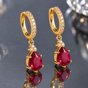 May include: A pair of gold hoop earrings featuring a row of small clear stones along the front of the hoop. Each earring has a dangling teardrop-shaped red stone pendant accented by three tiny clear stones at the top of the setting.