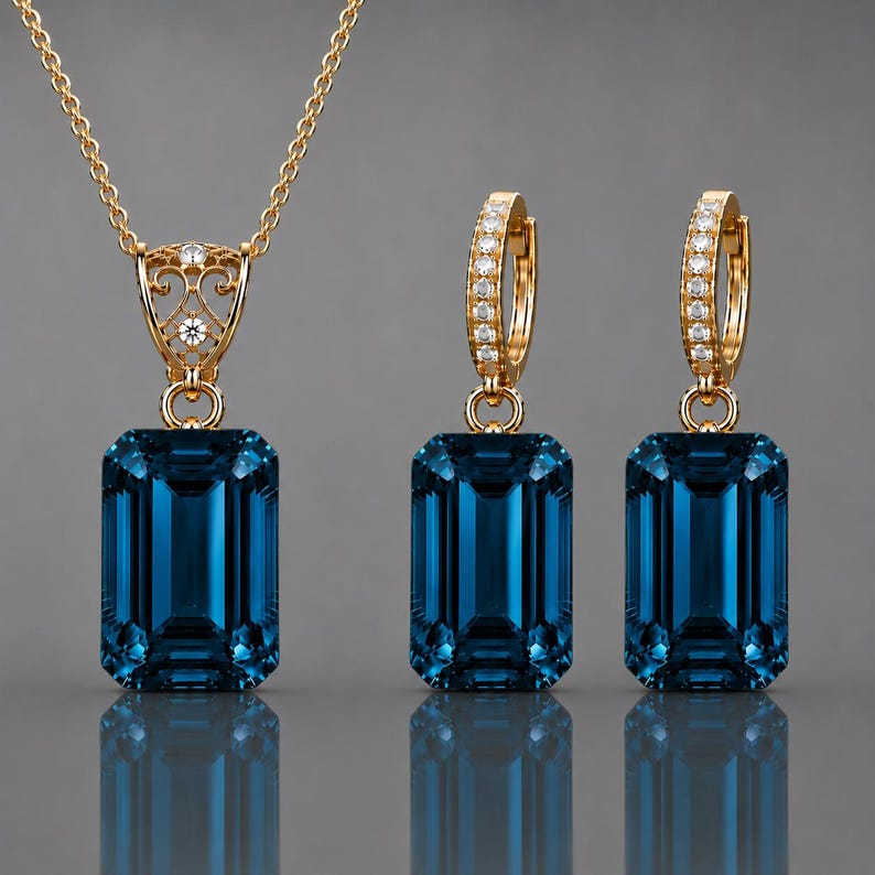 May include: A gold jewelry set consisting of a pendant necklace and two matching hoop earrings. Each piece features a large, emerald-cut, deep blue gemstone suspended from a gold setting adorned with small, clear, sparkling accent stones.
