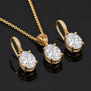 May include: A gold jewelry set consisting of a necklace and two matching earrings. Each piece features a large, oval-cut clear stone held in a gold prong setting, with smaller clear stones embedded along the earring hooks and pendant bail.