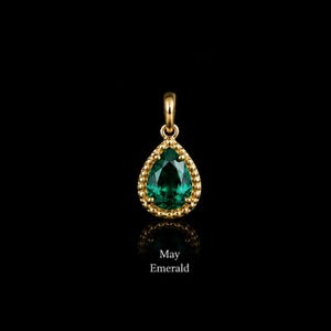 May include: A gold pendant featuring a pear-shaped green emerald gemstone set within a beaded gold frame. The pendant includes a small bail at the top for attachment. Below the pendant, the text May Emerald is printed in white lettering.