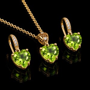 May include: A gold jewellery set consisting of a necklace and two earrings. Each piece features a heart-shaped, faceted lime green gemstone set in gold, with small clear stones embedded along the earring hooks and the necklace bail.