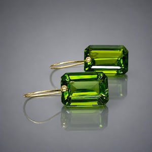 May include: A pair of elegant earrings featuring large, rectangular, vibrant green gemstones. The gemstones are set in gold-colored metal with a small, round accent. The earrings have a simple, curved hook design.