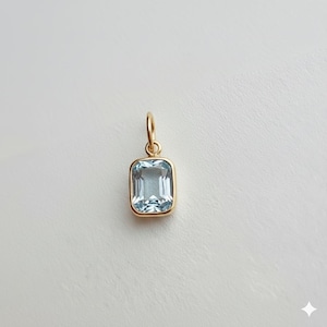 May include: A rectangular, light blue gemstone pendant with a gold-coloured bezel and loop. The pendant is approximately 1.3 cm in length and 0.8 cm in width. The gemstone has a clear, transparent appearance.