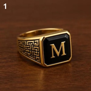 May include: A gold-toned signet ring featuring a square black inlay with a prominent gold letter M centred on the face. The sides of the ring band are decorated with an intricate, recessed geometric Greek key pattern in a dark, oxidised finish.