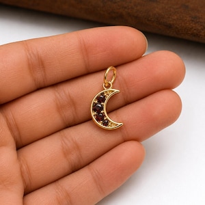 Pode incluir: Um pingente de lua crescente dourada com uma argola para uma corrente. A lua é incrustada com várias pedras preciosas pequenas, redondas e vermelho escuro. O pingente é segurado em uma mão.