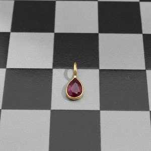Pode incluir: Um pingente de ouro com uma pedra preciosa vermelha escura em forma de gota. A pedra preciosa é colocada em uma moldura de ouro e tem um pequeno laço para uma corrente. O pingente é exibido em um fundo quadriculado preto e cinza.