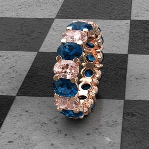 Puede incluir: Anillo de eternidad de oro rosa con piedras preciosas ovales azules y rosas alternas. El anillo se encuentra sobre un fondo a cuadros en blanco y negro, resaltando el diseño y el contraste de color. Una joya.