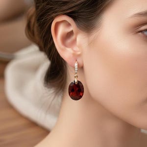 Peut inclure: Boucles d'oreilles pendantes dorées avec une grosse pierre précieuse ovale rouge foncé. Les boucles d'oreilles ont une petite section décorative avec des pierres claires près du lobe de l'oreille.