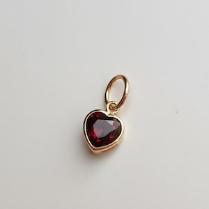 May include: A gold-coloured heart-shaped pendant with a deep red gemstone. The pendant is designed to be worn as a necklace charm or earring. The gemstone has a faceted surface. The pendant is suspended from a small loop.