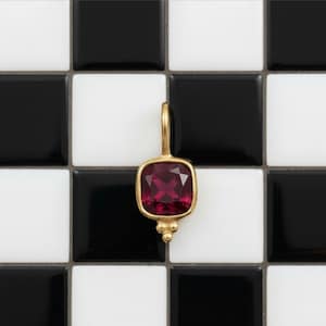 Könnte beinhalten: Ein goldener Anhänger mit einem quadratischen, tiefroten Edelstein. Der Anhänger hat oben eine Schlaufe und unten eine Ansammlung von drei kleinen goldenen Perlen. Der Hintergrund ist ein schwarz-weißes Schachbrettmuster.