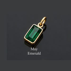 May include: A gold pendant featuring a rectangular emerald-cut green gemstone. The pendant has a small circular bail at the top for attachment. Below the pendant, the text "May Emerald" is printed in white, sans-serif capital and lowercase letters.