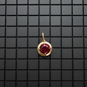 May include: A circular gold pendant featuring a single round-cut deep red gemstone set in the center. The pendant has a small gold bail at the top for attachment to a chain. The design is simple, highlighting the contrast between the gold and red.