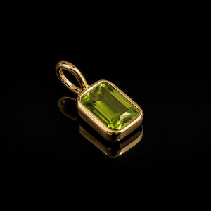 Op de afbeelding: Een gouden hanger met een rechthoekige peridoot edelsteen in smaragdgeslepen vorm, gezet in een gladde, gepolijste gouden zetting. De hanger is voorzien van een klein, rond gouden oogje aan de bovenzijde en heeft een levendige limoengroene kleur.