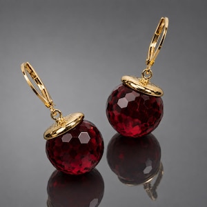 Puede incluir: Un par de pendientes colgantes con cuentas de cristal esféricas de color rojo oscuro facetado, suspendidas de cierres de palanca en tono dorado pulido. Las cuentas redondas tienen una superficie texturizada y facetada con una tapa dorada.
