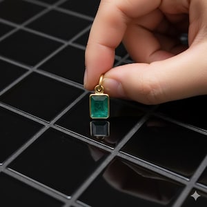 Puede incluir: Un colgante de oro con una piedra preciosa de esmeralda rectangular. La esmeralda es de color verde vibrante y está engastada en un bisel de oro. El colgante está sostenido por una mano.