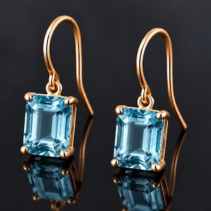 Könnte beinhalten: Ein Paar goldene Ohrhänger mit rechteckigen, im Smaragdschliff geschliffenen hellblauen Edelsteinen. Jeder Stein wird von vier Goldkrappen gehalten und hängt an einem geschwungenen Goldhaken, was ein klassisches Design mit poliertem Finish ergibt.
