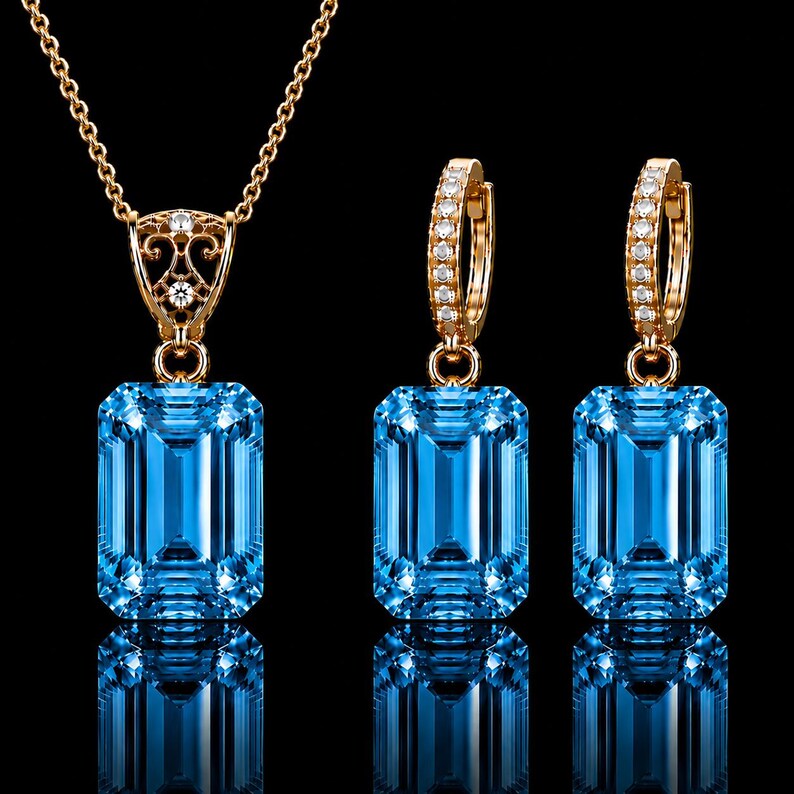 May include: A jewelry set consisting of a necklace and two hoop earrings, each featuring a large, rectangular, emerald-cut blue gemstone. The gold-toned settings are accented with small, clear, round-cut stones on the pendant bail and hoops.