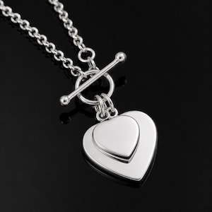 Puede incluir: Un collar de tono plateado con cierre de barra y un colgante de doble corazón. El colgante consta de dos dijes en forma de corazón lisos y pulidos superpuestos, suspendidos de un anillo circular conectado a una cadena de eslabones.