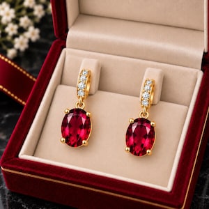 May include: A pair of gold drop earrings featuring oval-cut deep red gemstones suspended from hoops encrusted with small clear stones. The earrings are presented in a rectangular jewellery box with a dark red velvet exterior and beige interior.