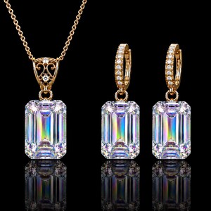 May include: A gold jewelry set consisting of a necklace and two hoop earrings. Each piece features a large, emerald-cut clear stone with iridescent light reflections. The necklace pendant and earring hoops are accented with small, clear pav&eacute; stones.