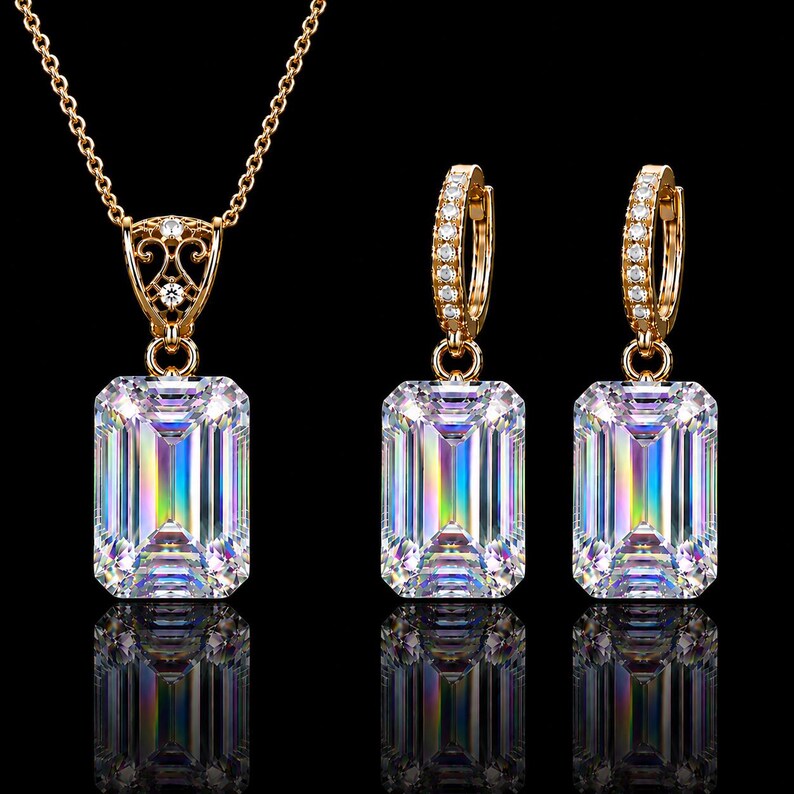 May include: A gold jewelry set consisting of a necklace and two hoop earrings. Each piece features a large, emerald-cut clear stone with iridescent light reflections. The necklace pendant and earring hoops are accented with small, clear pav&eacute; stones.