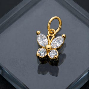 May include: A gold-colored butterfly charm with clear, sparkling crystal accents. The charm features a loop for attaching to a necklace or bracelet. The butterfly's wings are made of marquise-cut crystals, and the body has a round crystal.