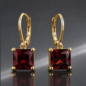 May include: A pair of gold leverback earrings featuring square-cut, deep red garnet gemstones. Each earring has a polished gold setting with four prongs securing the faceted square stone, suspended from a curved gold leverback closure mechanism.