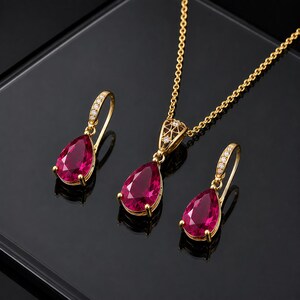 May include: A gold jewellery set consisting of a necklace and two drop earrings. Each piece features a pear-shaped, deep pink gemstone set in gold. The earring hooks and the necklace bail are accented with small, clear, round-cut white stones.