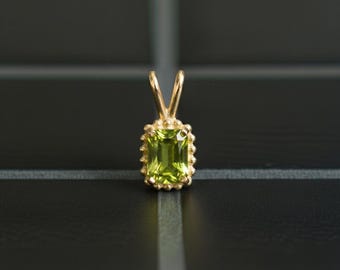May include: A gold pendant featuring a rectangular, emerald-cut lime green gemstone. The stone is set in a gold frame with a beaded edge and a triangular bail at the top for a chain. The design is symmetrical and highlights the faceted green gem.