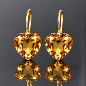 May include: A pair of gold-toned drop earrings featuring faceted, heart-shaped amber-colored crystals. Each earring is suspended from a curved gold-toned wire hook, showcasing a brilliant, multi-faceted surface that reflects light with clarity.